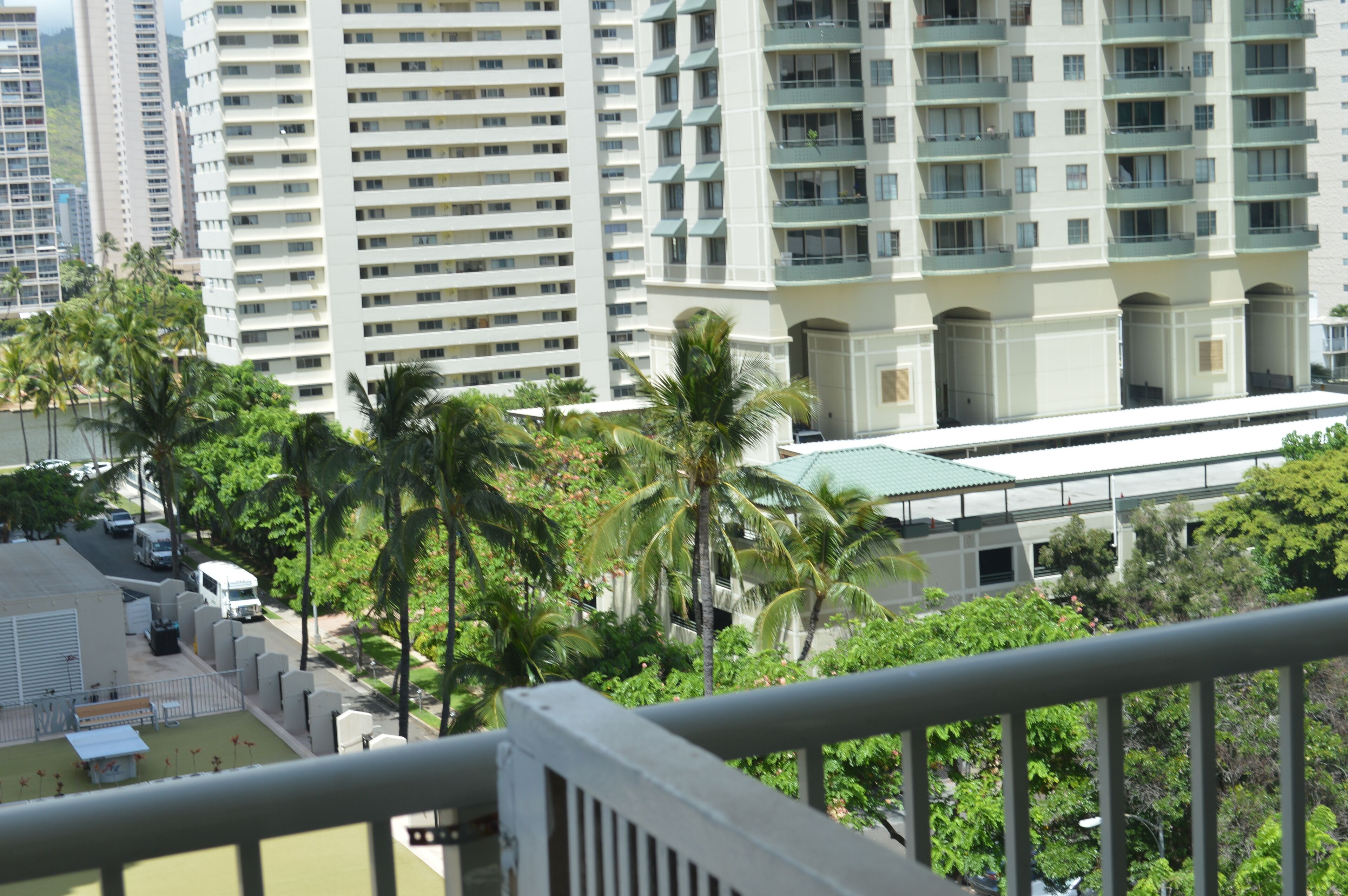 A high-rise building with multiple balconies, surrounded by palm trees and a road with vehicles.