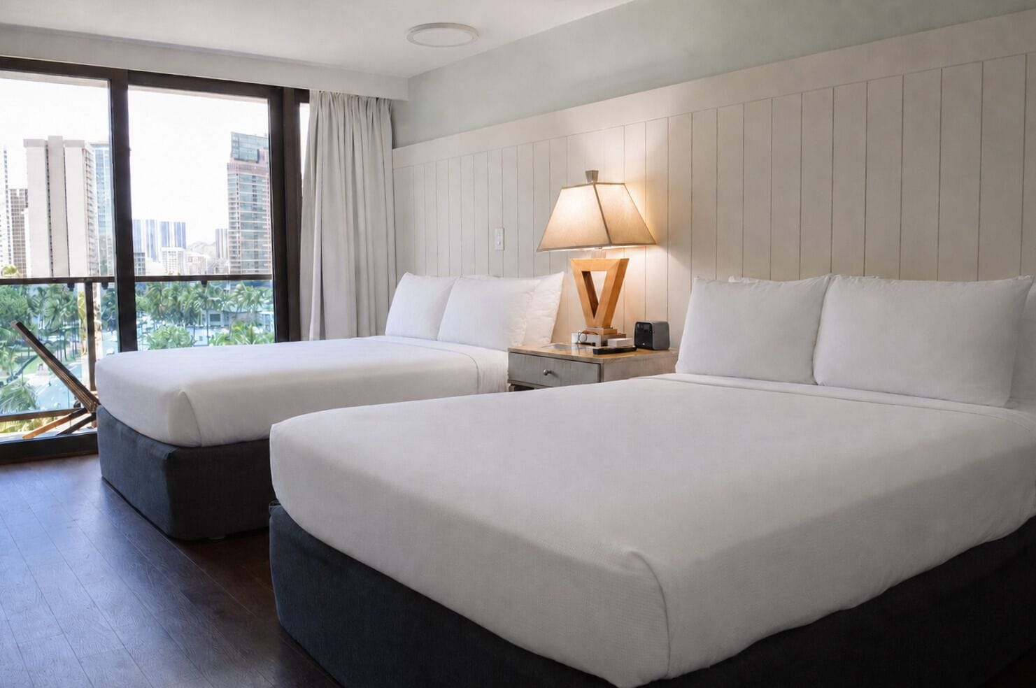 Una camera d'albergo con due letti, un comodino, una lampada e vista su palme e palazzi.