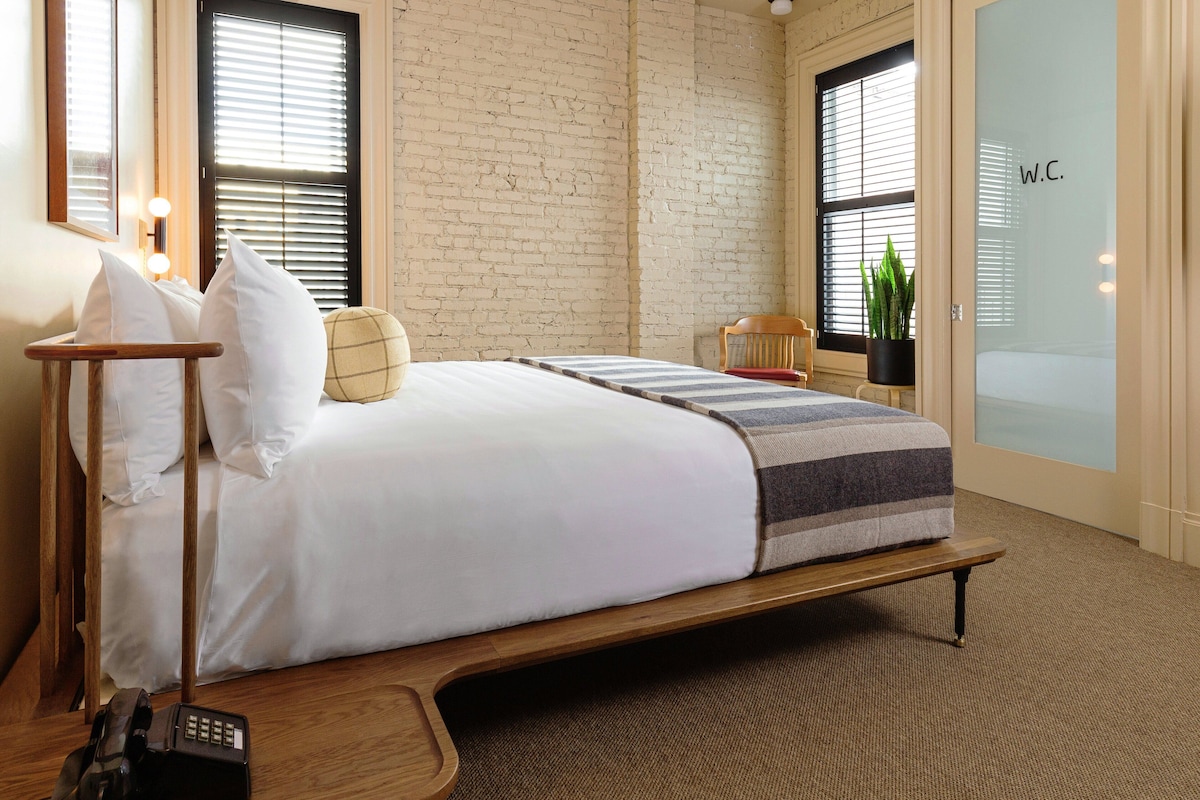 A modern hotel room with a brick wall, a large bed with white bedding, a wooden bedside table, and a bedside lamp.