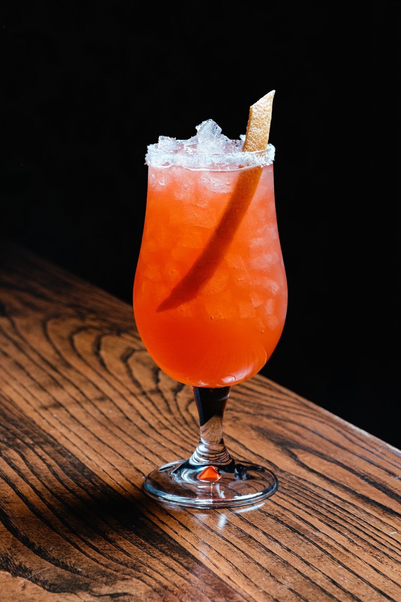 A cocktail in a tall glass with a salted rim and a wafer stick, placed on a wooden surface.
