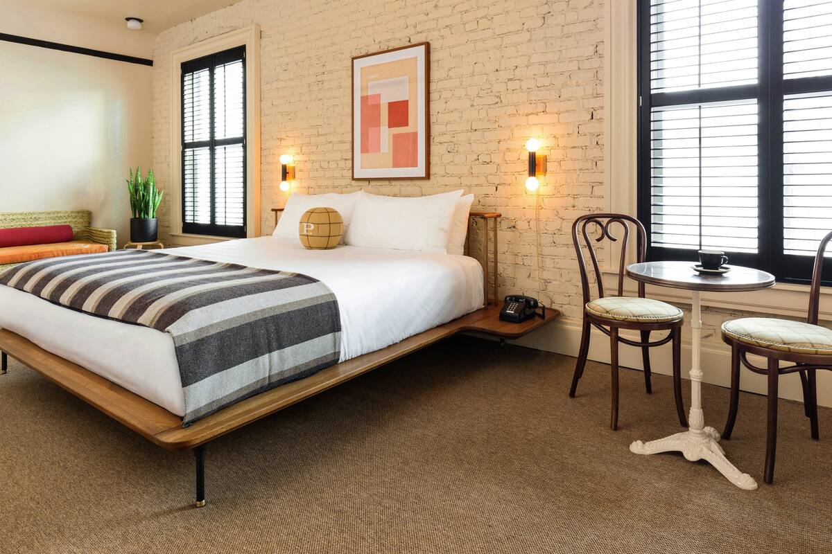 A hotel room with a bed, two chairs, a small table, a potted plant, and a framed picture on the wall.