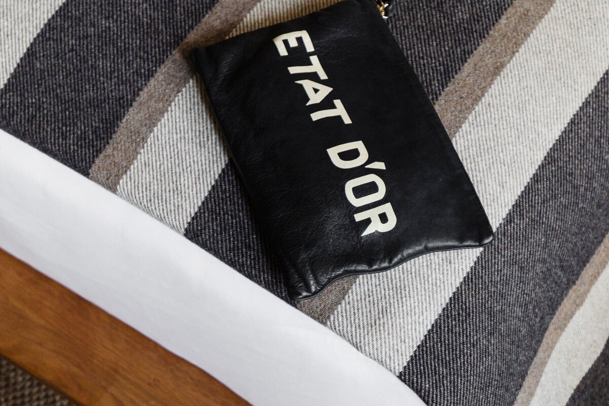 Un sac à main noir orné de l’inscription « ETAT D’OR », des chaussures blanches et un coussin rayé sur une chaise en bois.