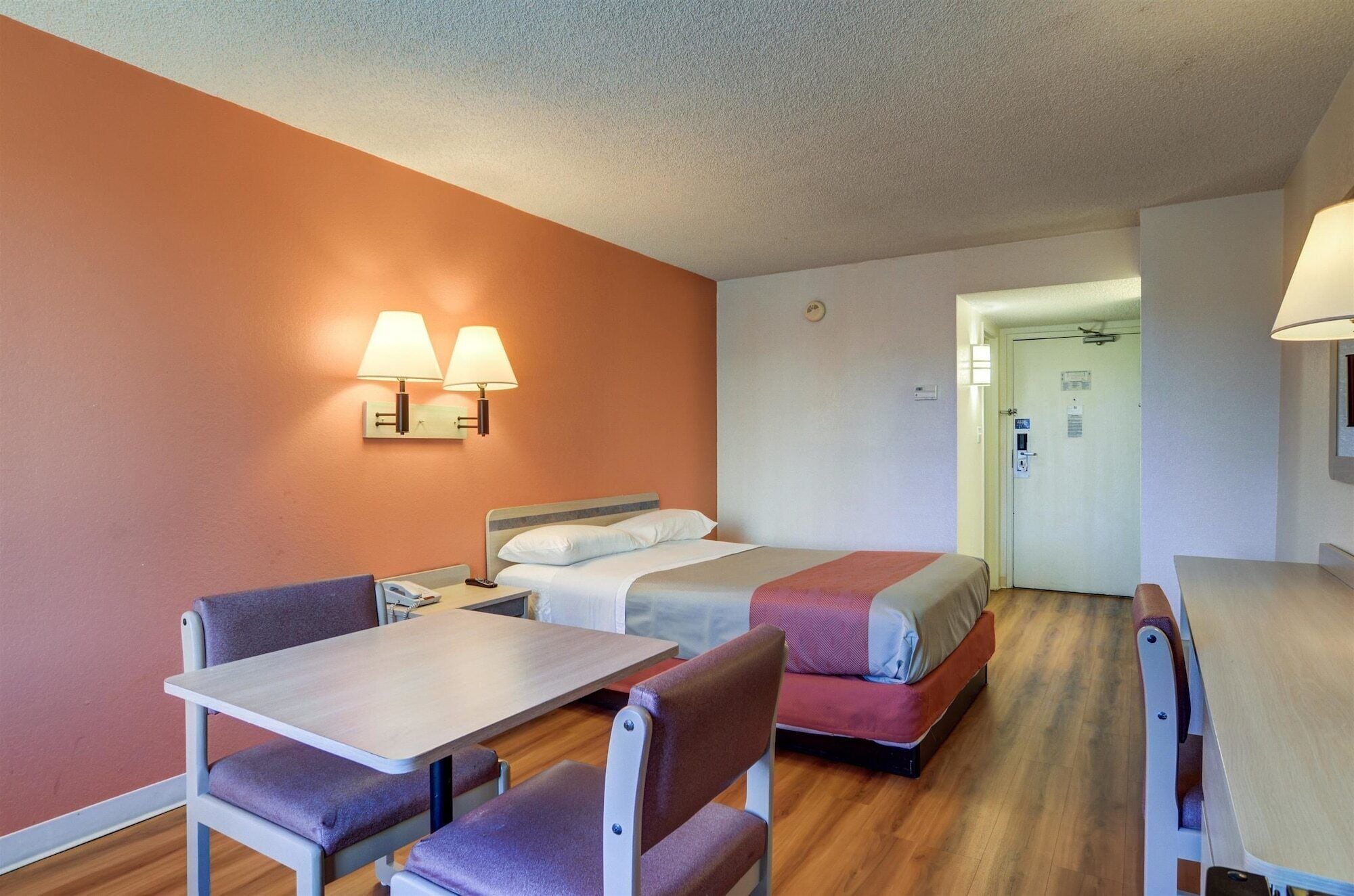Une chambre d’hôtel avec un lit, deux tables de chevet, un bureau et une chaise. Il y a deux lampes fixées au mur et une porte avec une serrure.