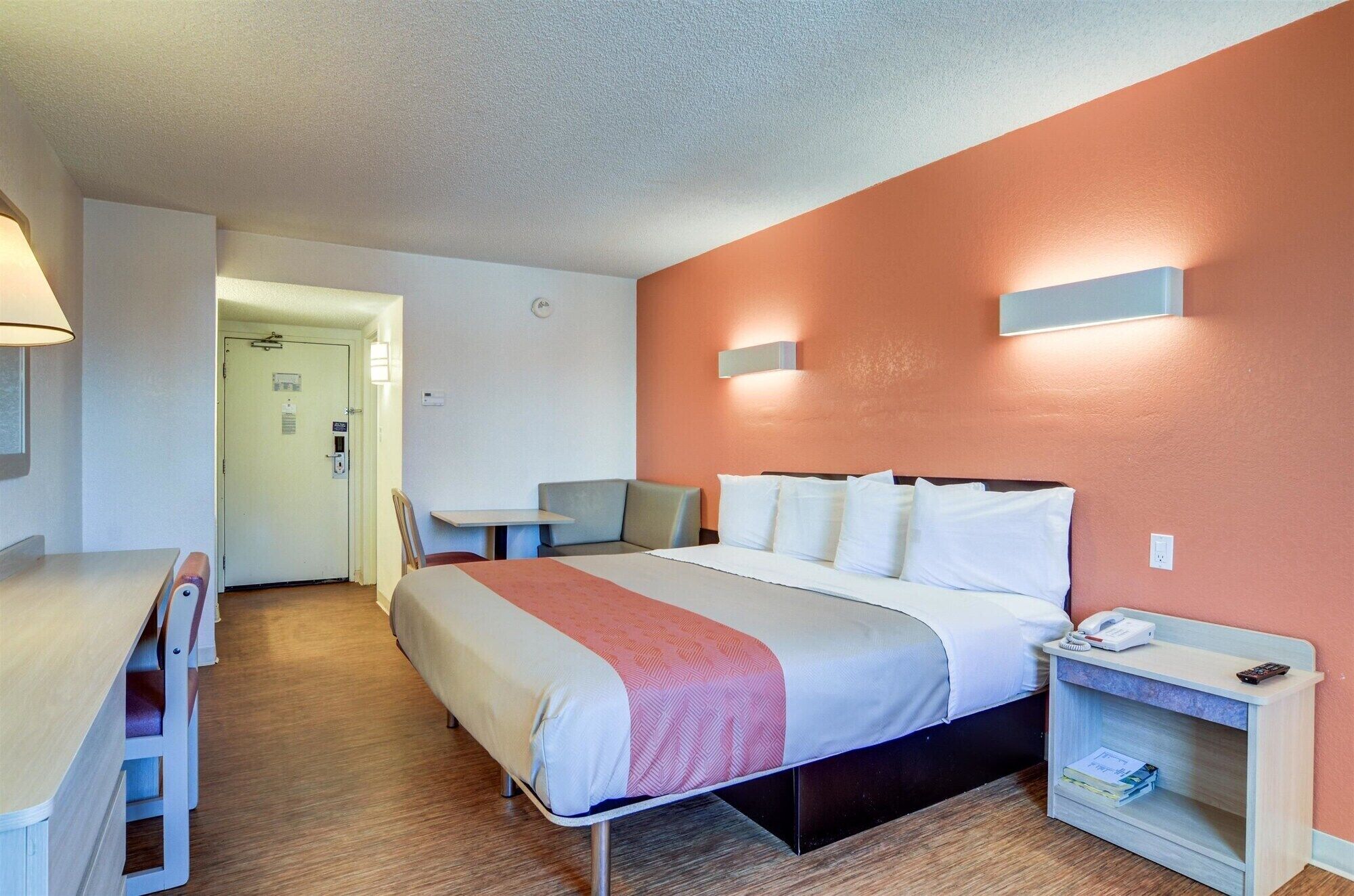 A hotel room with a bed, bedside table, chair, and a desk with a lamp.