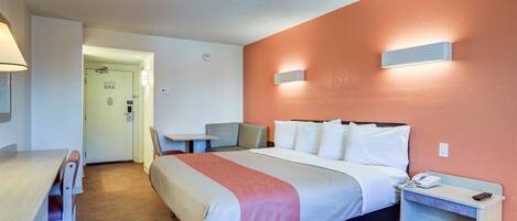 A hotel room with a bed, bedside table, chair, and a desk with a lamp.