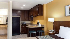 Habitación de hotel con cama, escritorio con lámpara, y kitchenette equipada con refrigerador, microondas y cafetera.