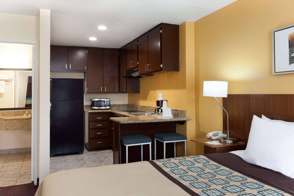 Habitación de hotel con cama, escritorio con lámpara, y kitchenette equipada con refrigerador, microondas y cafetera.