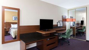 Habitación de hotel con un escritorio de madera, televisor de pantalla plana, silla de oficina verde, espejo y cama con alfombra púrpura.