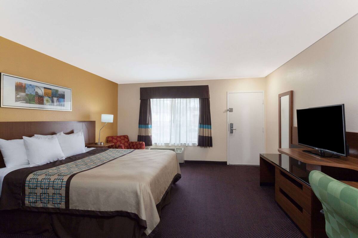 Habitación de hotel con cama, escritorio con televisor de pantalla plana, una silla, ventana con cortinas y puerta.