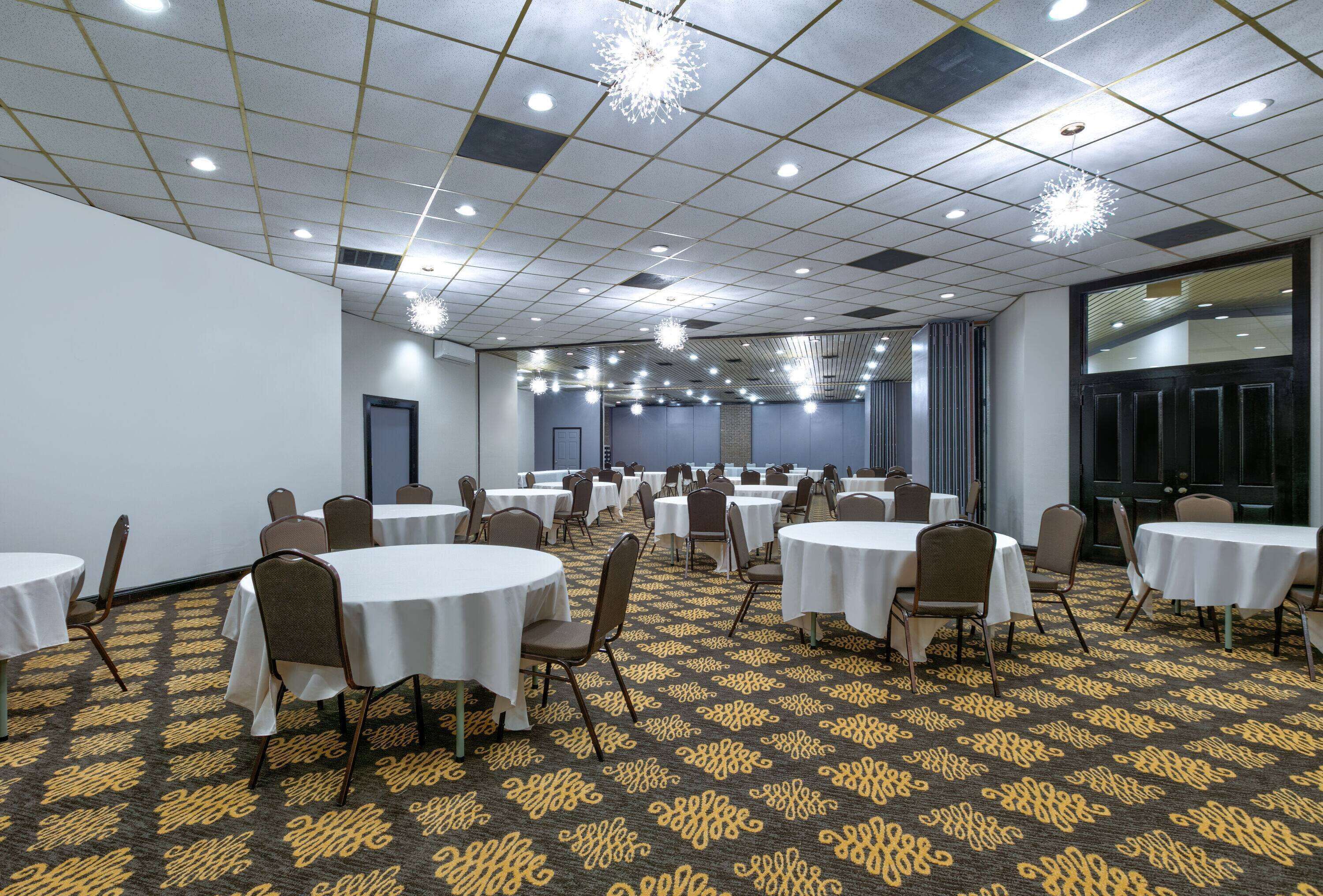 A conference room with multiple round tables and chairs, a patterned carpet, and a grid ceiling with recessed lighting.