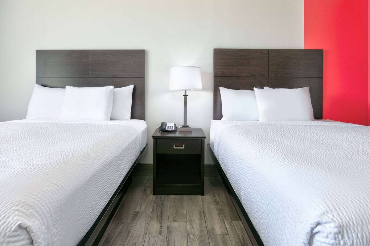 Habitación de hotel con dos camas, una mesita de noche con lámpara y una pared de acento roja.