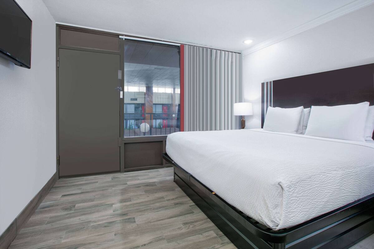 Habitación de hotel con cama, televisor, ventana con persianas y puerta.