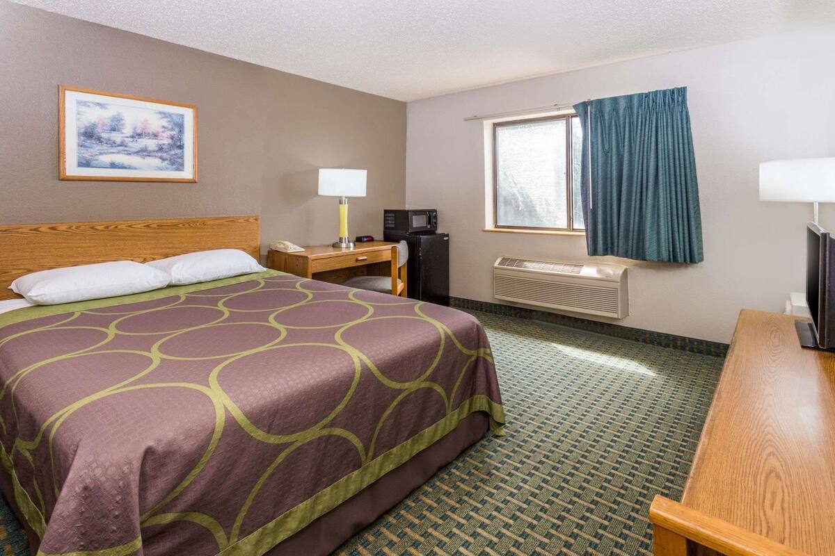 Una camera d'albergo con un letto, una scrivania, una televisione, una finestra con tende, un quadro appeso al muro e un condizionatore d'aria.