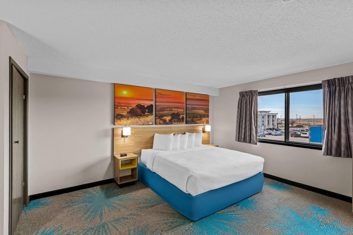 Une chambre d’hôtel avec un lit, une table de chevet, une lampe fixée au mur, une fenêtre avec des rideaux et une vue sur la ville.
