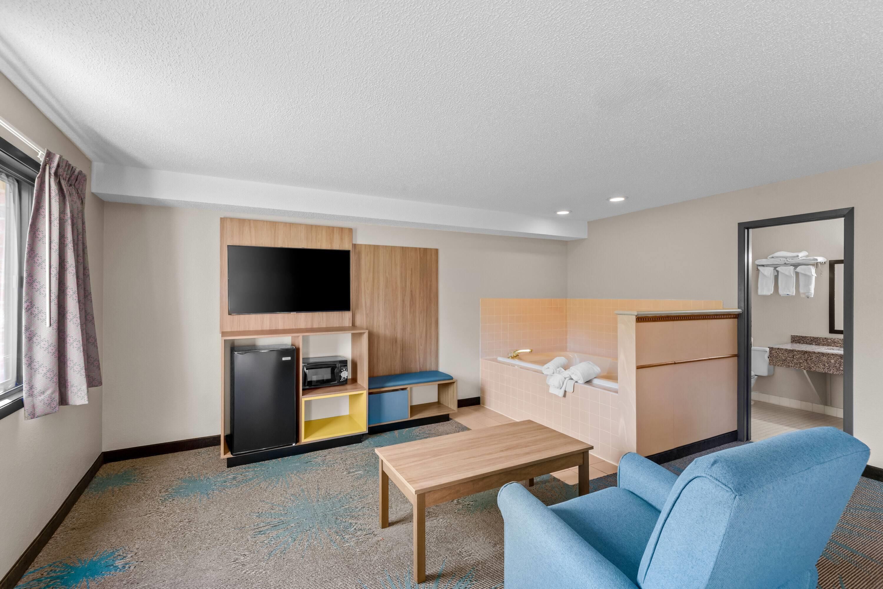 Habitación de hotel compacta con kitchenette, televisor de pantalla plana, un pequeño rincón de descanso y baño con espejo y toallero.