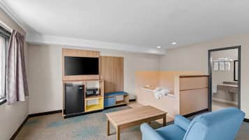 Habitación de hotel compacta con kitchenette, televisor de pantalla plana, un pequeño rincón de descanso y baño con espejo y toallero.