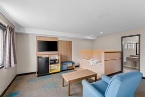A compact hotel room with a kitchenette, a flat-screen TV, a small seating area, and a bathroom with a mirror and towel rack. - Days Inn & Suites by Wyndham Des Moines Airport (Des Moines)