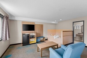 Habitación de hotel compacta con kitchenette, televisor de pantalla plana, un pequeño rincón de descanso y baño con espejo y toallero.