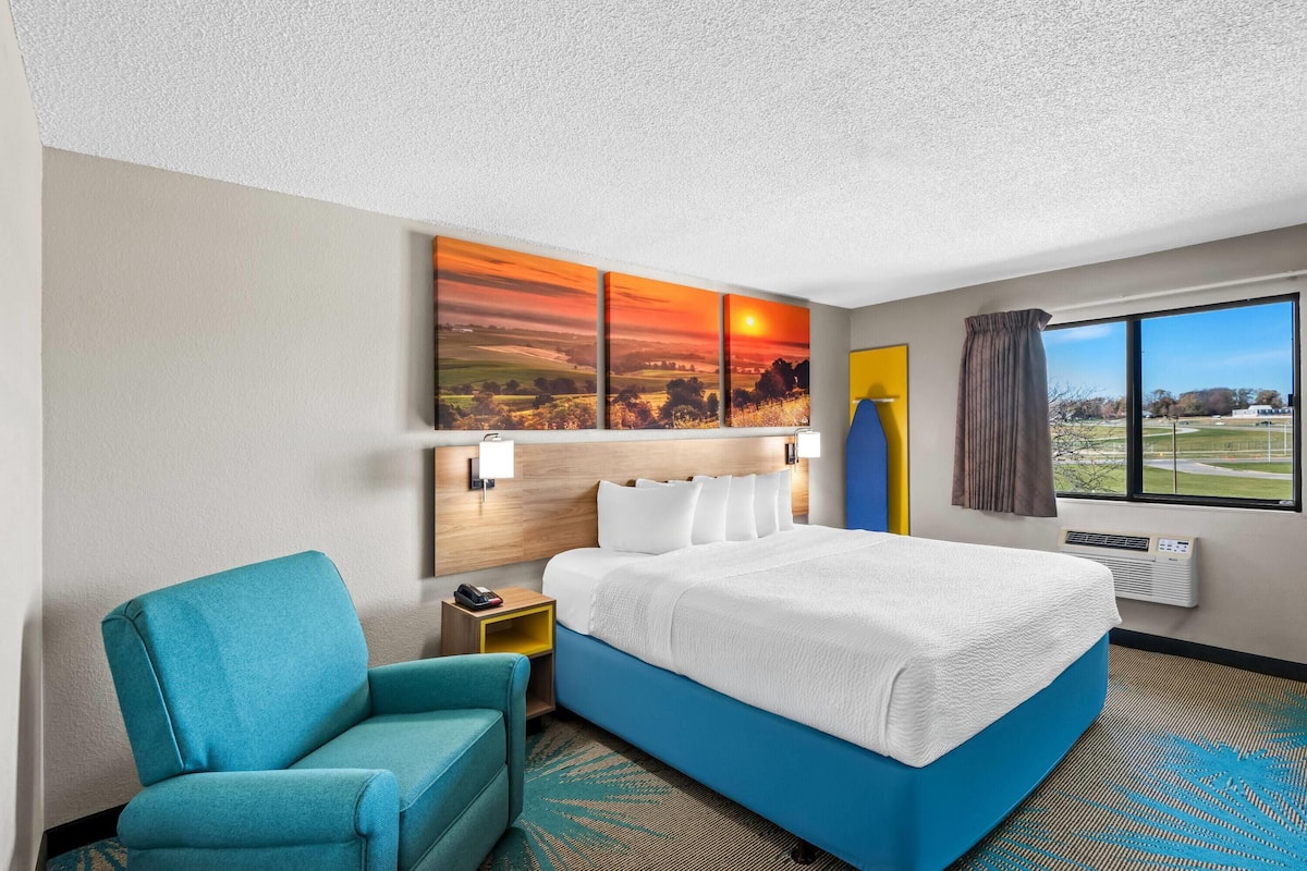Une chambre d’hôtel avec un grand lit, un mur d’accent bleu, un tableau représentant un coucher de soleil, une vue sur un terrain de golf et un fauteuil turquoise.