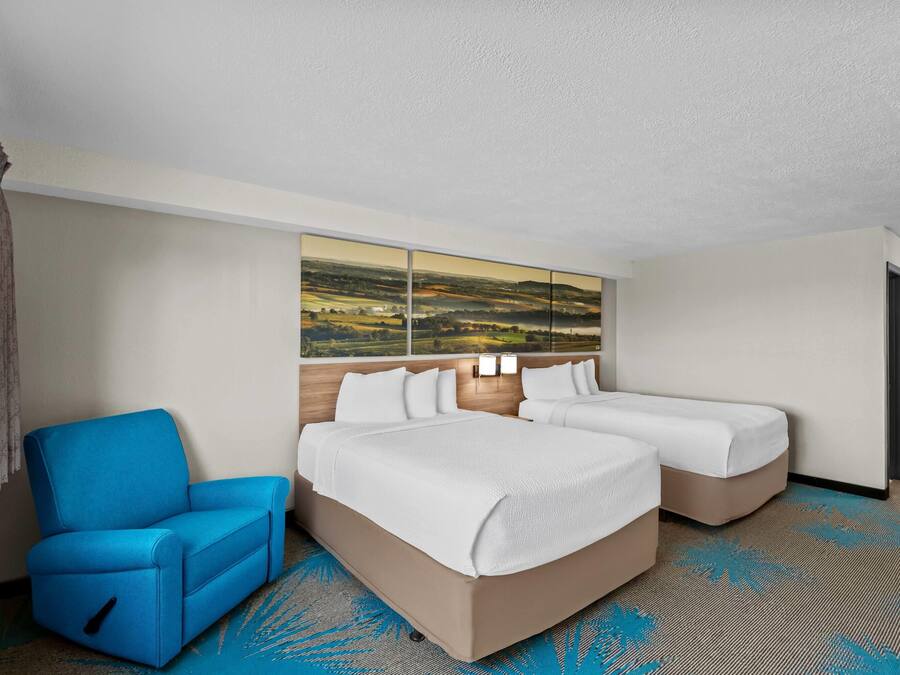 A hotel room with two beds, a blue armchair, a large landscape painting on the wall, and a patterned carpet.