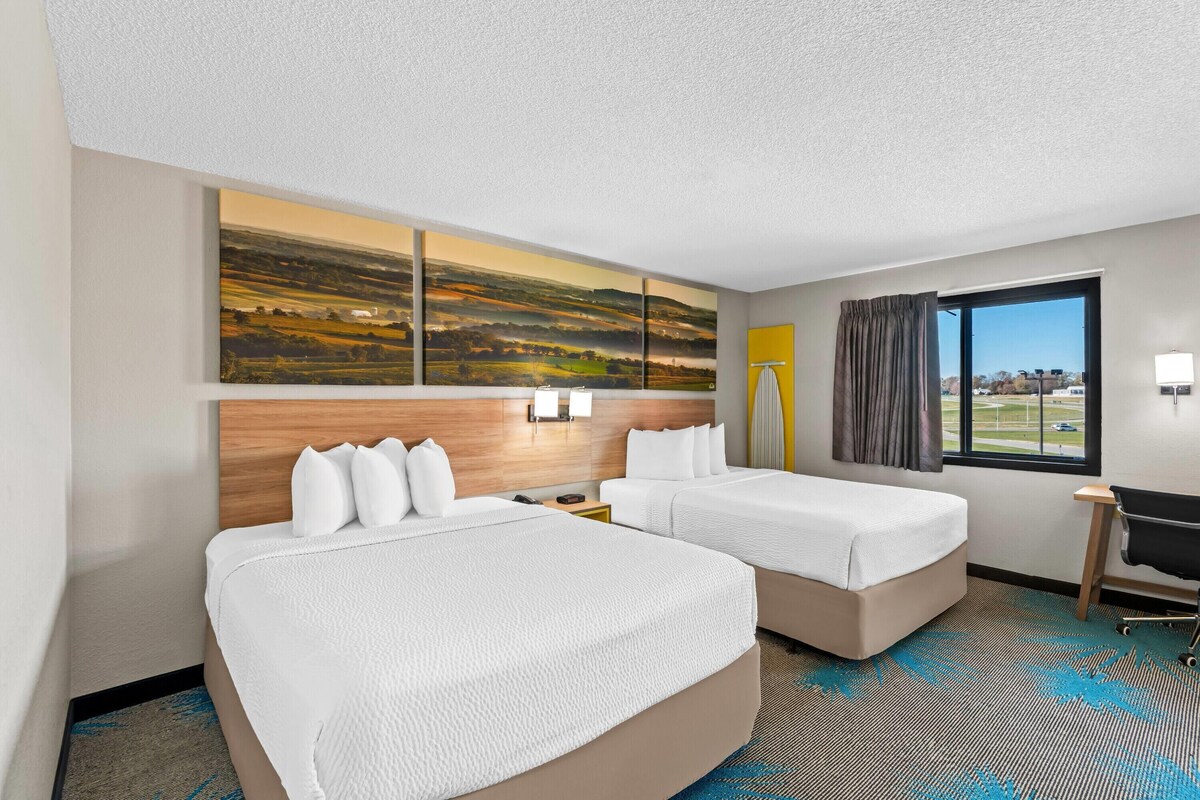 A hotel room with two beds, a large landscape painting, a desk with a chair, and a view of the outdoors through a window.