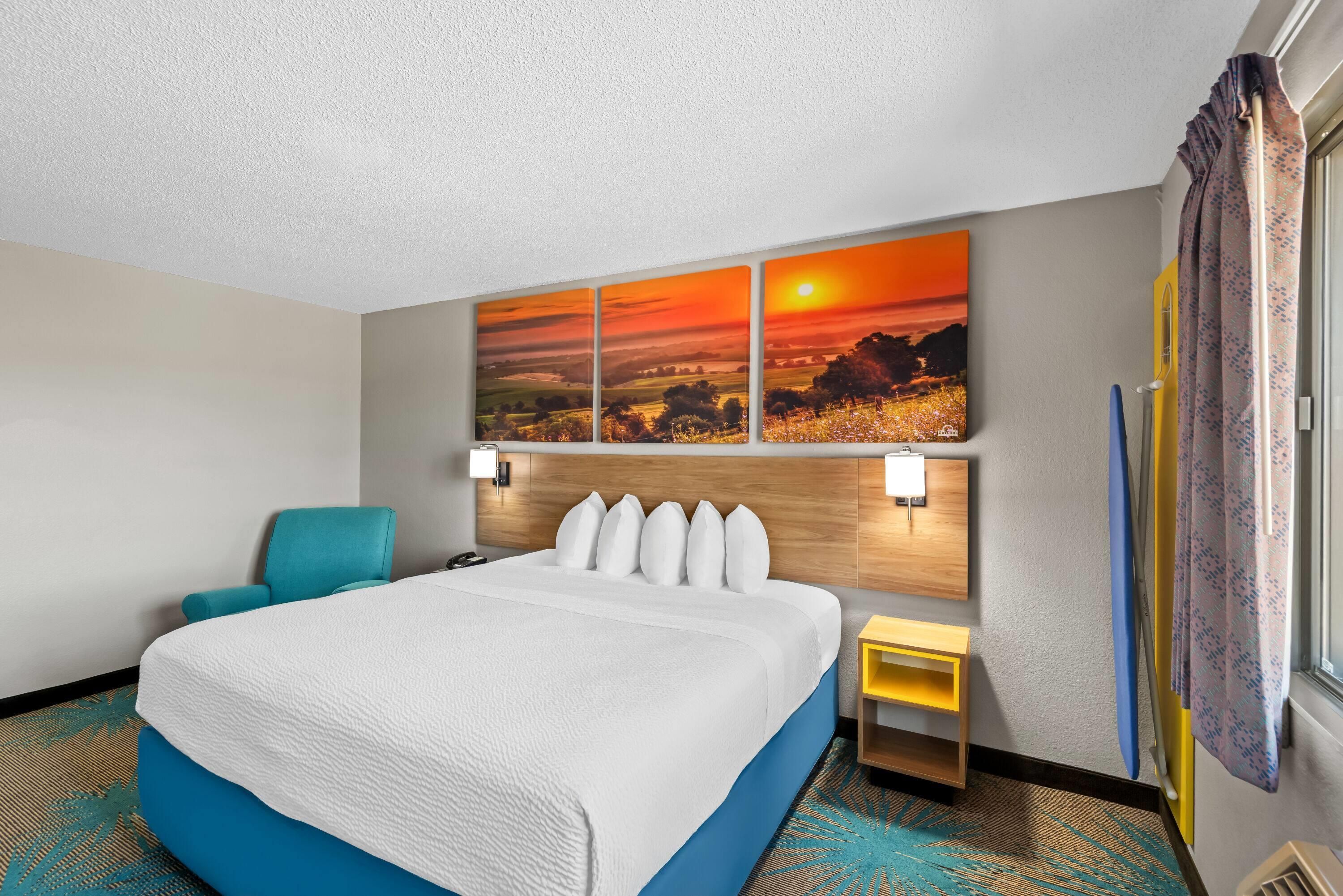 Une chambre d’hôtel avec un grand lit, une tête de lit dotée de lampes, un tableau représentant un coucher de soleil, une petite table et une fenêtre avec des rideaux.