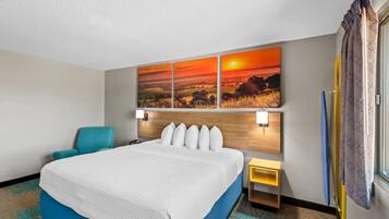 Une chambre d’hôtel avec un grand lit, une tête de lit dotée de lampes, un tableau représentant un coucher de soleil, une petite table et une fenêtre avec des rideaux.