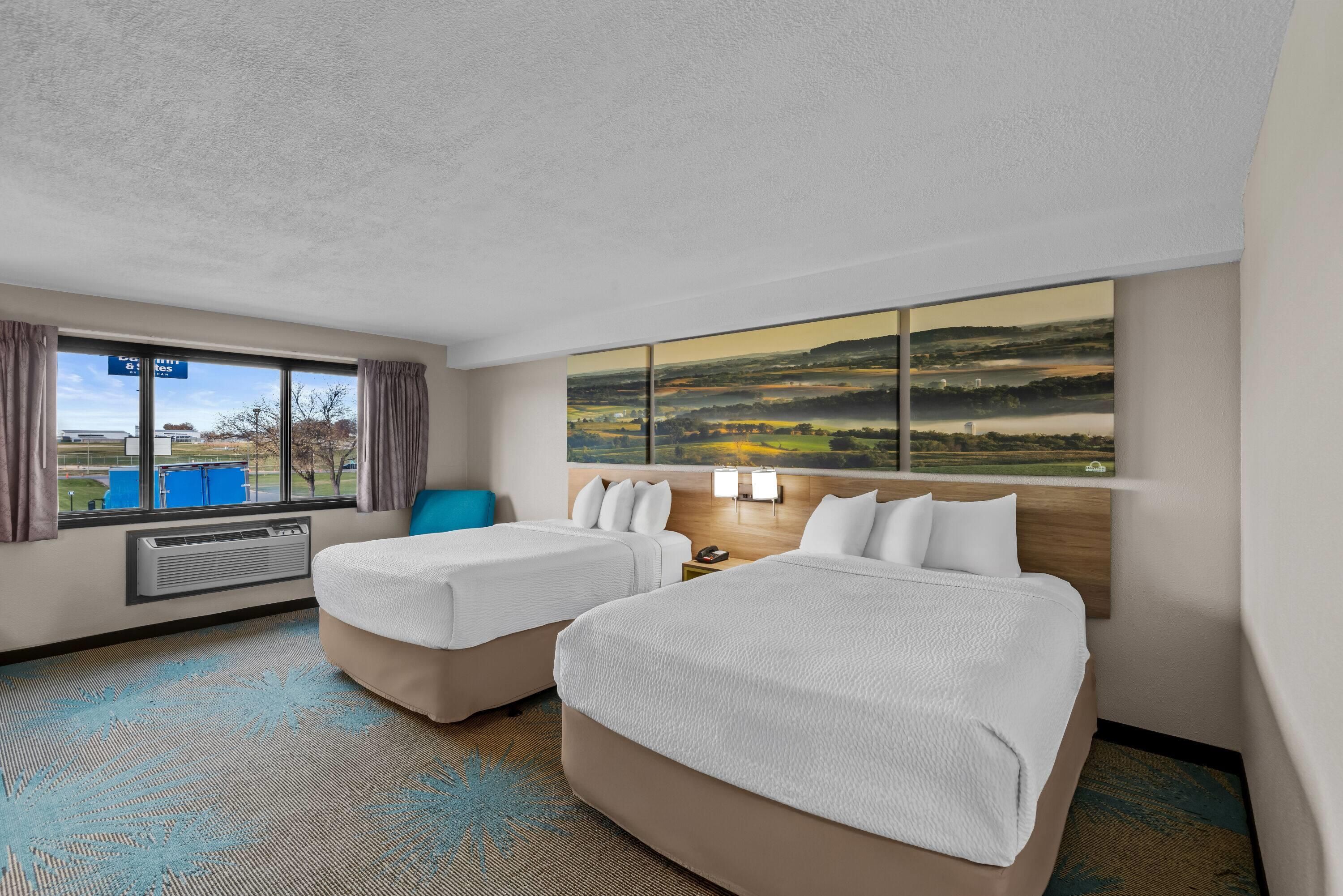 A hotel room with two beds, a large window with a countryside view, a wall-mounted air conditioner, and a blue accent pillow.
