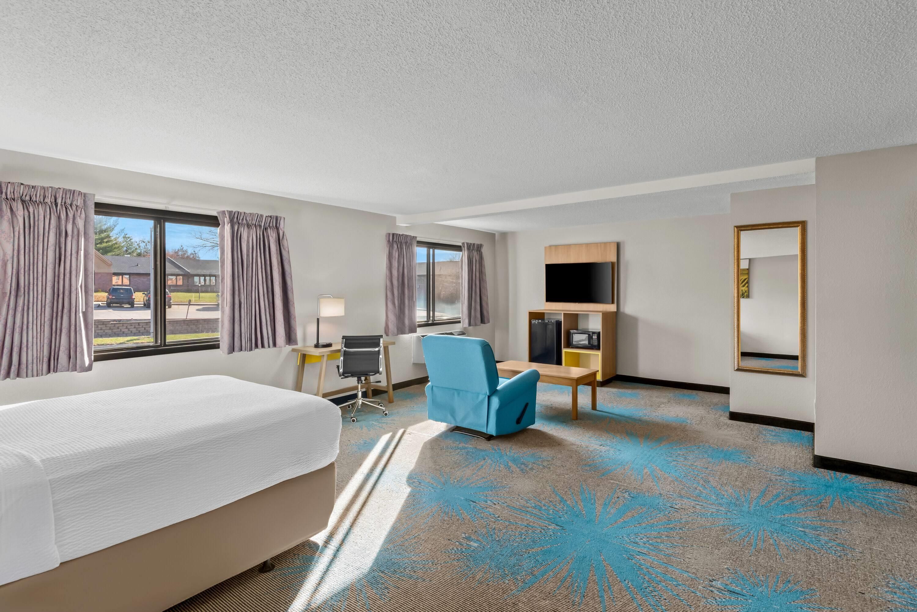 Habitación de hotel con cama, escritorio, silla, televisor y ventana grande con cortinas.