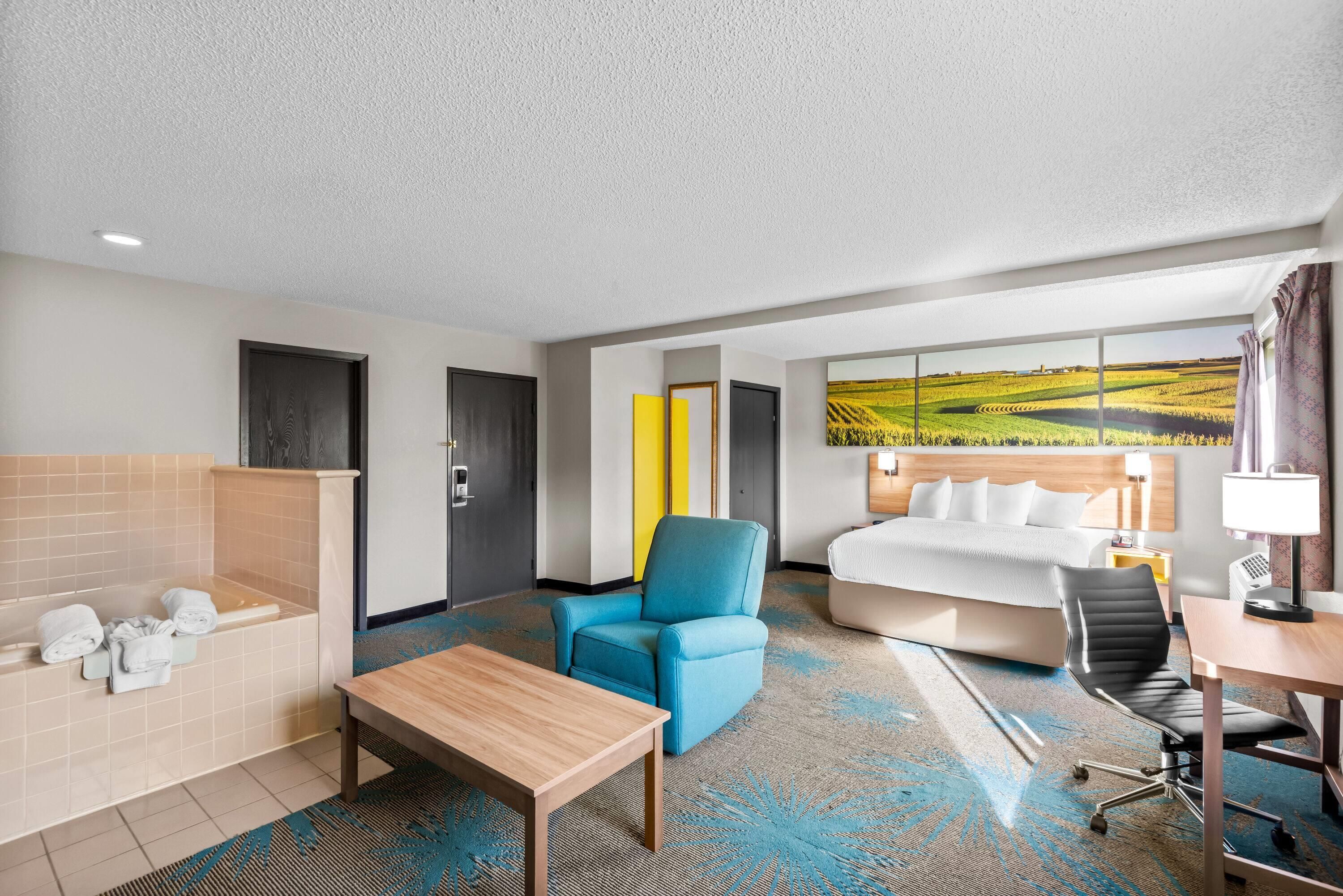 Habitación de hotel con una cama grande, un sillón azul, una mesa de madera, un escritorio con una silla, un baño con bañera y un ventanal con vistas a campos.