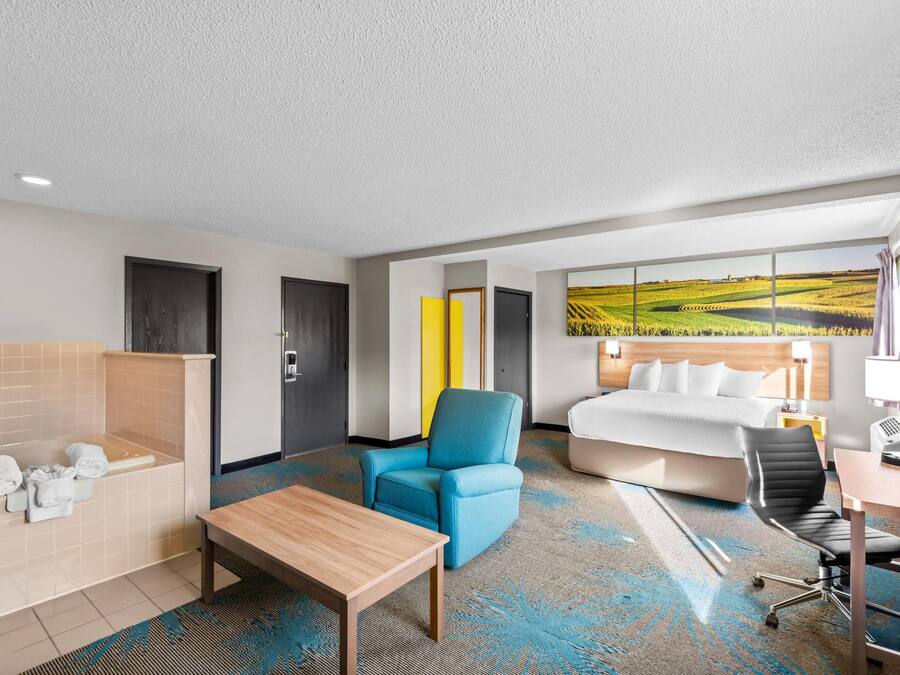 Habitación de hotel con una cama grande, un sillón azul, una mesa de madera, un escritorio con una silla, un baño con bañera y un ventanal con vistas a campos.
