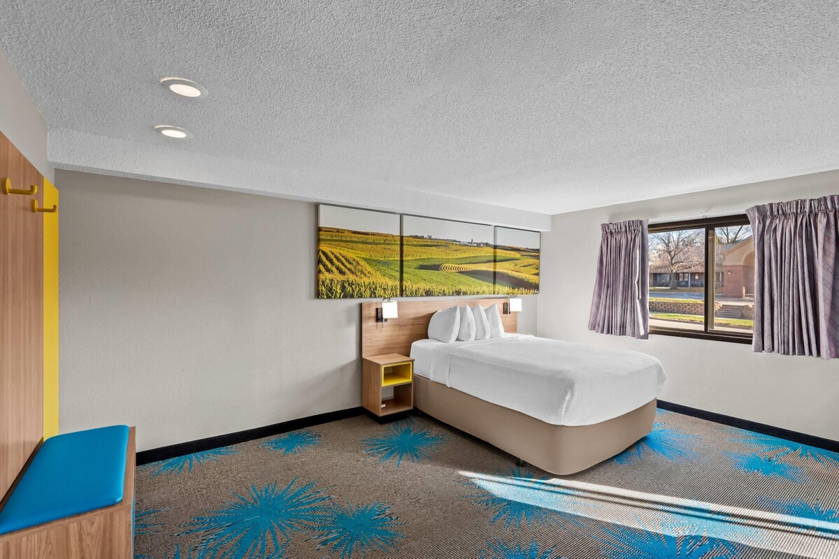 A hotel room with a bed, a nightstand, a desk with a chair, a large window with curtains, and a painting on the wall.