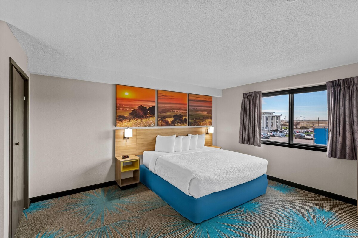 A hotel room with a bed, a nightstand, a wall-mounted lamp, a window with curtains, and a city view.