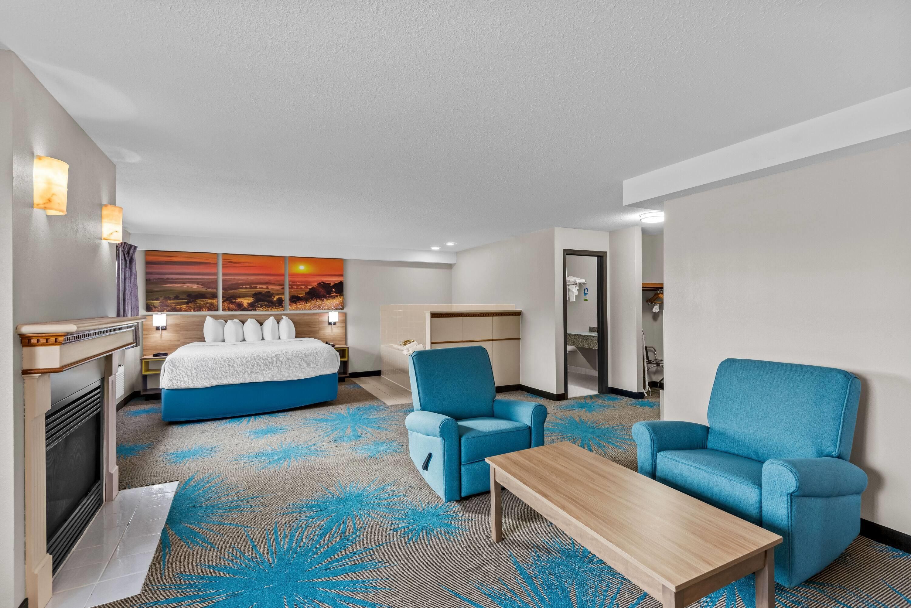 A hotel room with a bed, two armchairs, a coffee table, a fireplace, and a large window with a sunset view.