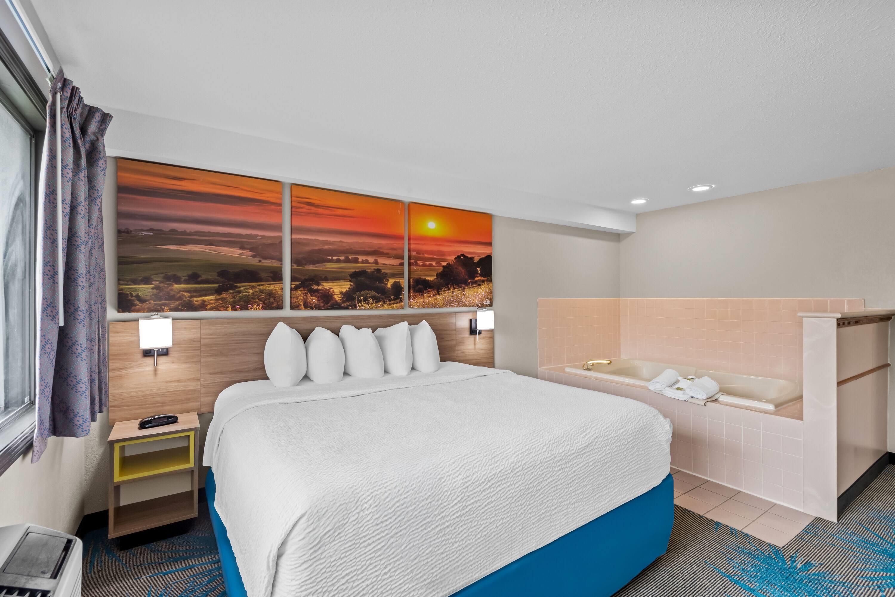 A hotel room with a large bed, a nightstand, a wall-mounted TV, a bathroom with a bathtub, and a scenic sunset painting on the wall.