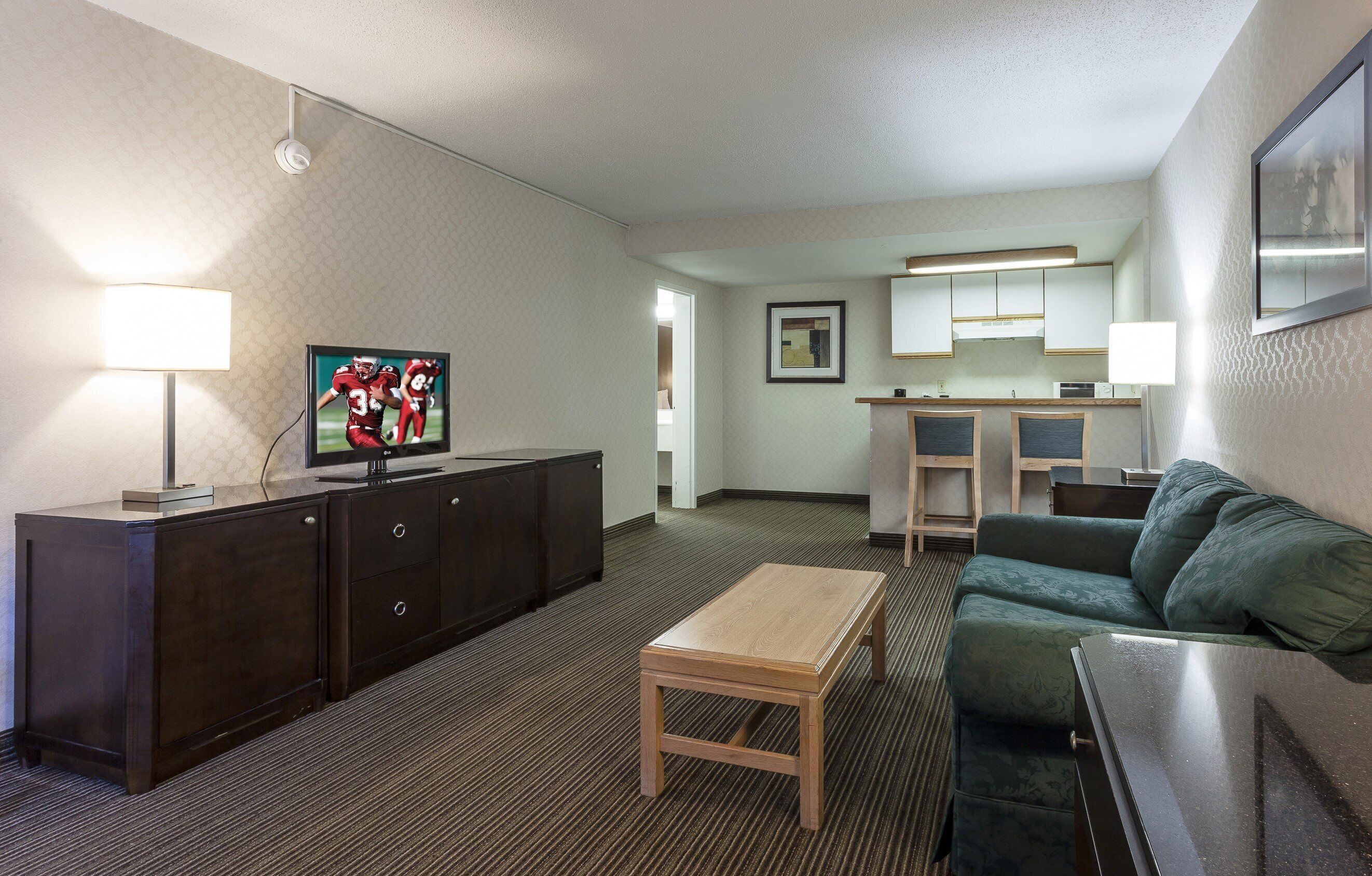 A hotel room with a television, a wooden table, a green sofa, a kitchenette, and a framed picture on the wall.