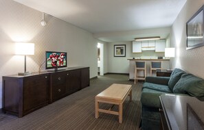 Habitación de hotel con televisión, mesa de madera, sofá verde, kitchenette y un cuadro en la pared.