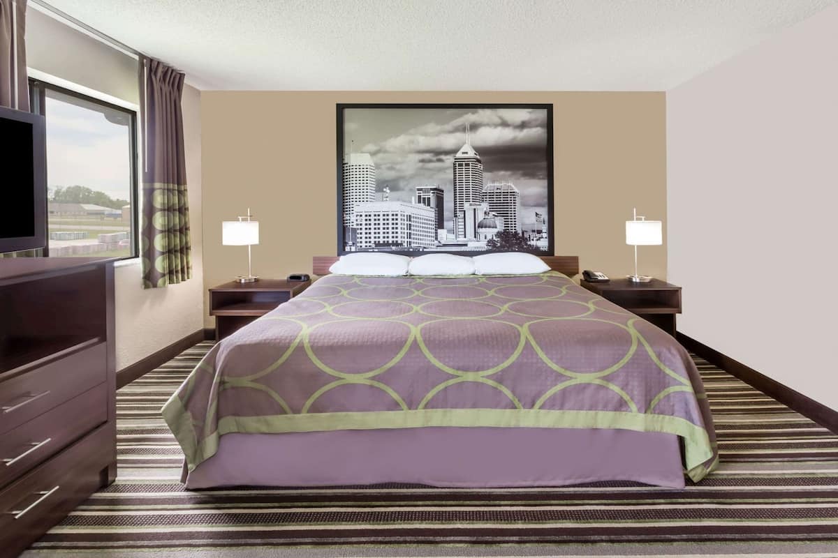 Un cuarto de hotel con una cama grande, dos mesitas de noche con lámparas, un aparador, una ventana con cortinas y un cuadro de un paisaje urbano en la pared.