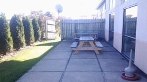 A patio with a wooden table and benches, a concrete floor, and a white wall with windows. - Super 8 by Wyndham Plymouth (Plymouth)