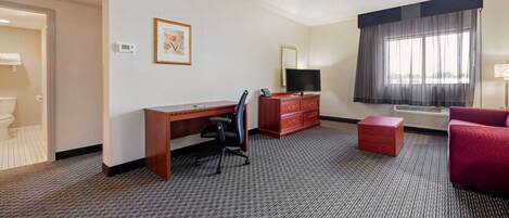 A hotel room with a desk, chair, television, dresser, and a red armchair.