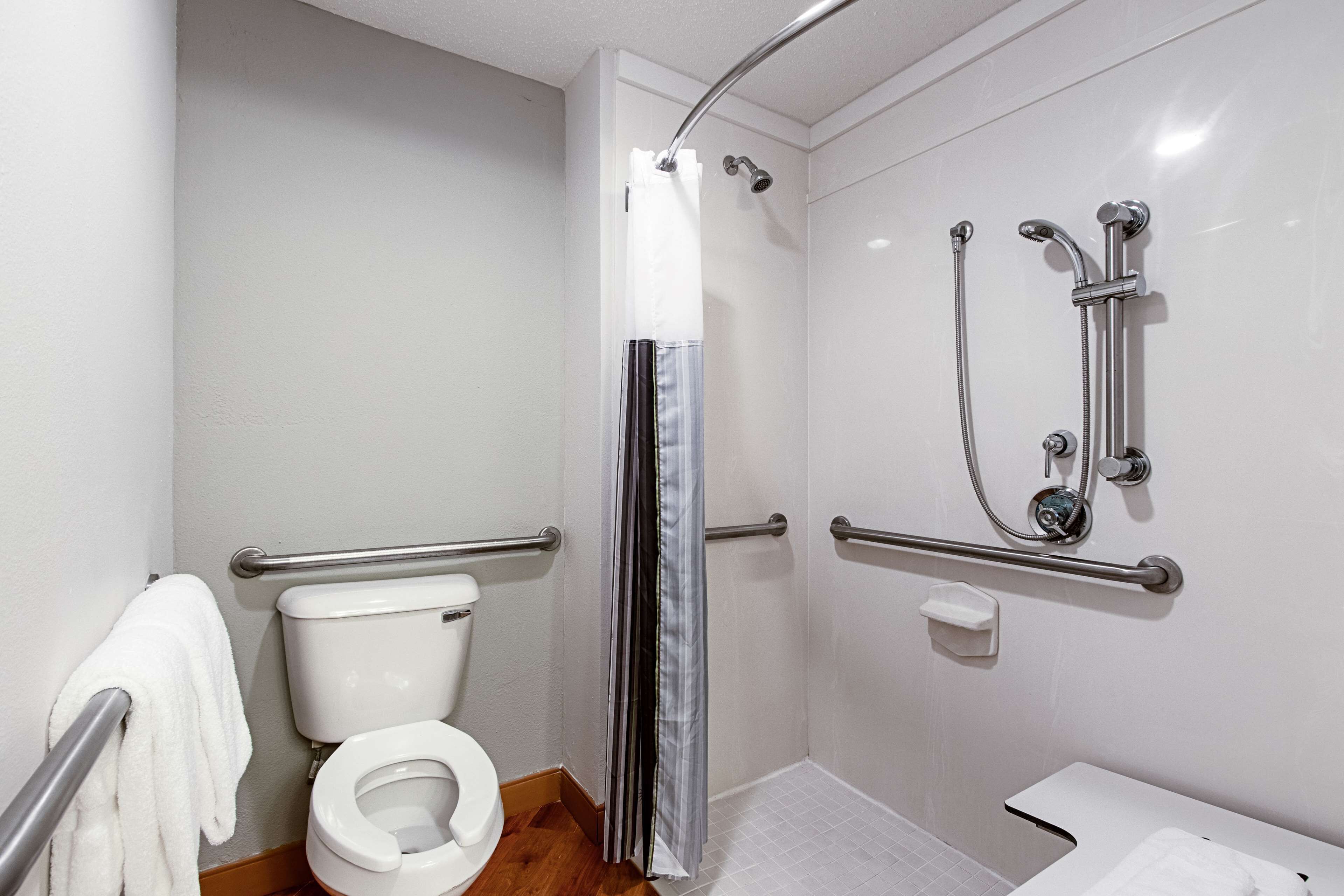 A bathroom with a white toilet, a shower with a handheld showerhead, grab bars, and a white towel on the floor.