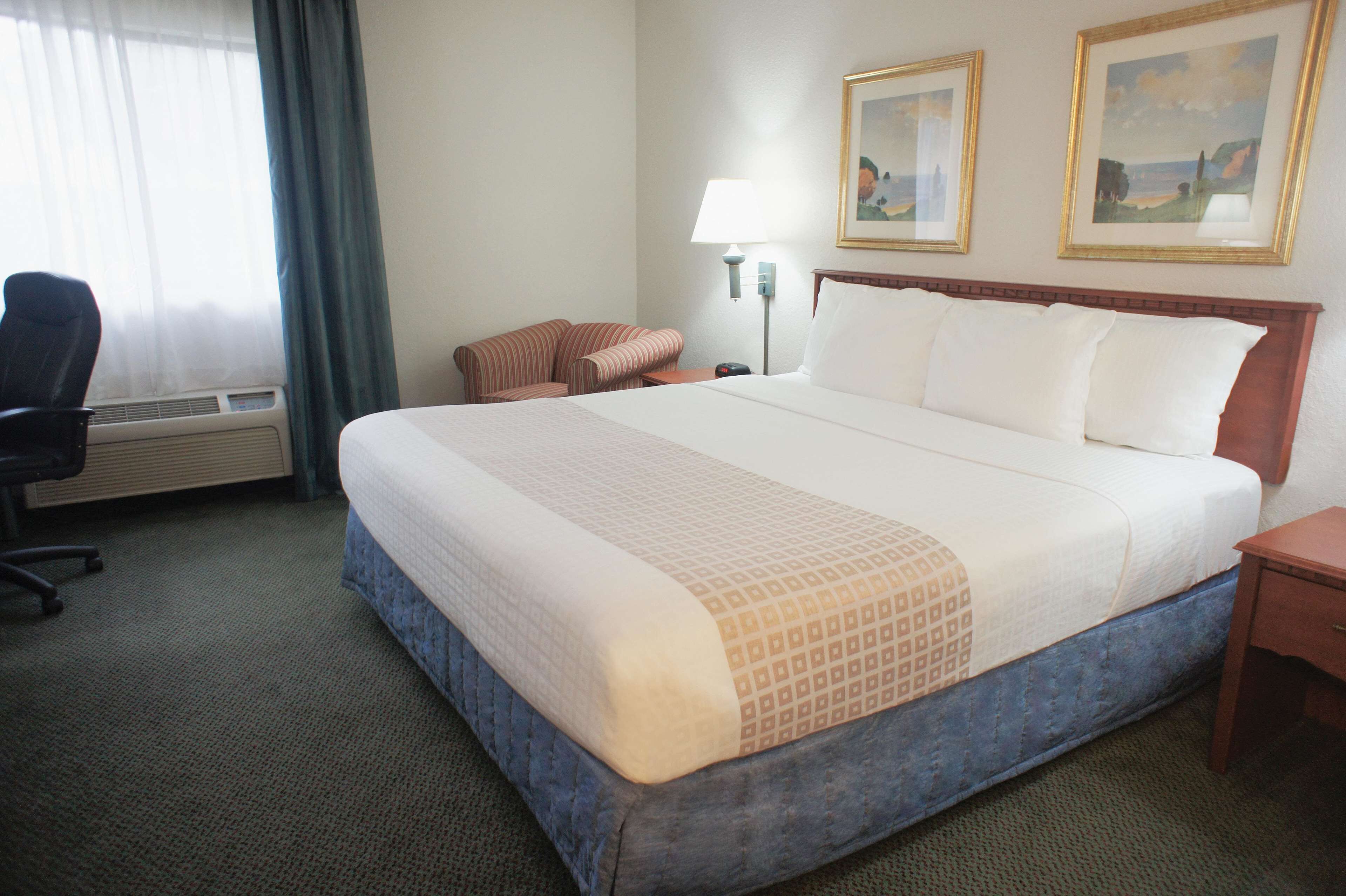 A hotel room with a large bed, a desk, a chair, a lamp, and two framed pictures on the wall.