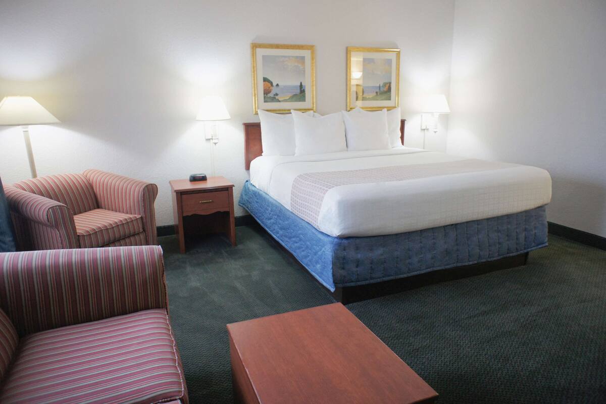 A hotel room with a bed, a striped armchair, a small wooden nightstand, and a night lamp.