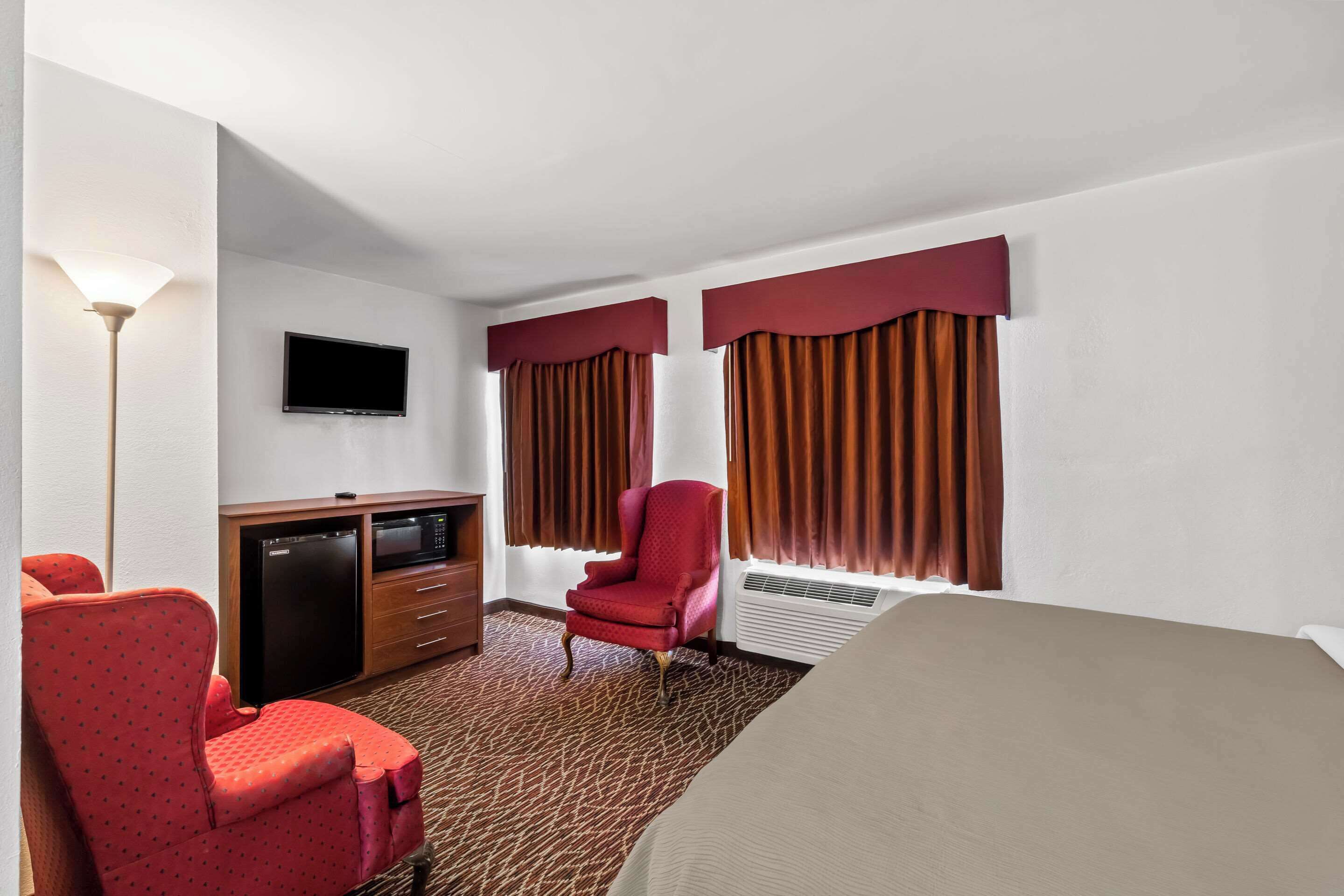 A hotel room with a bed, a red armchair, a television, a minibar, and a carpeted floor.