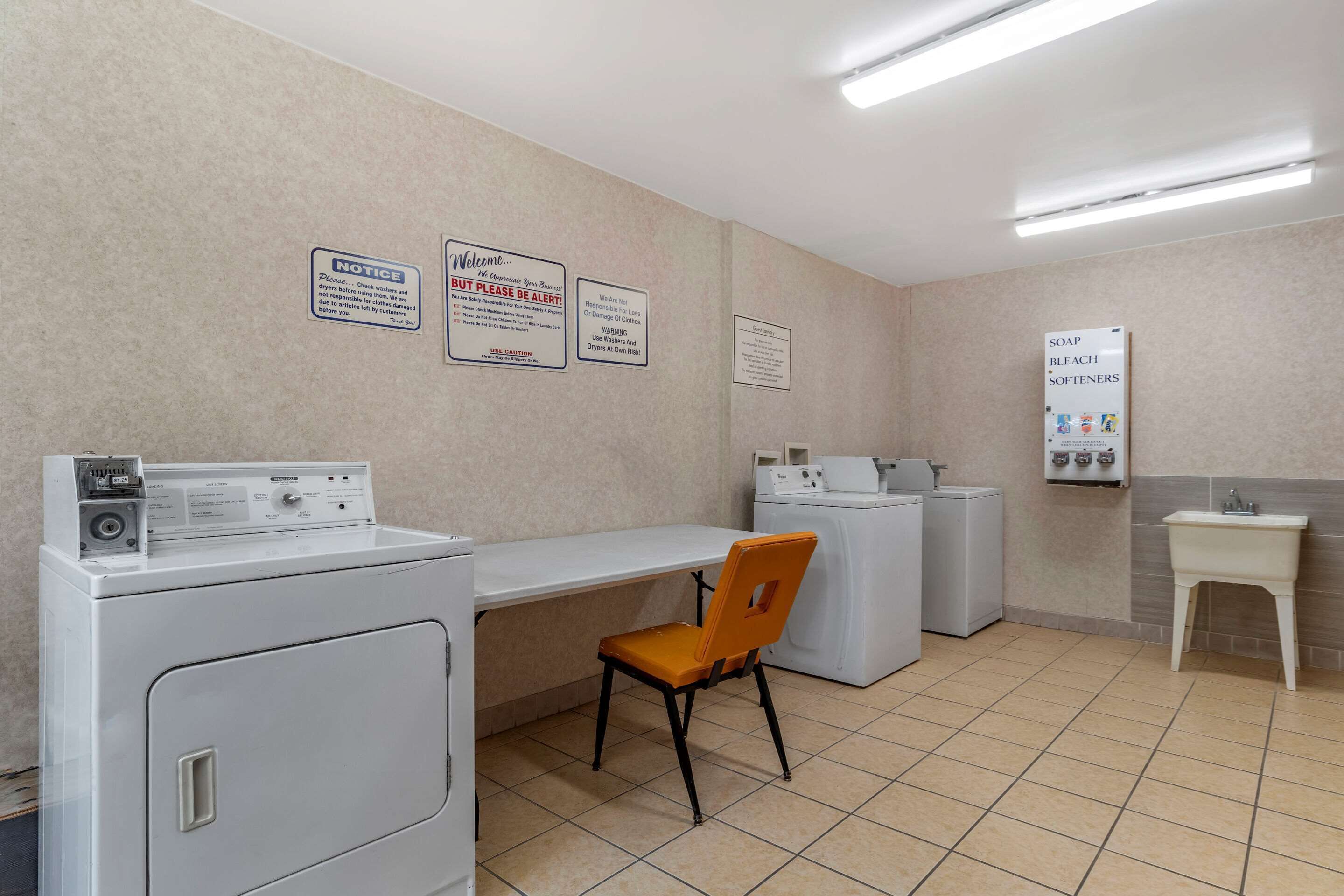 Una sala de lavandería con lavadora, secadora y una silla. Hay letreros en la pared y un lavabo con jabón y lejía.