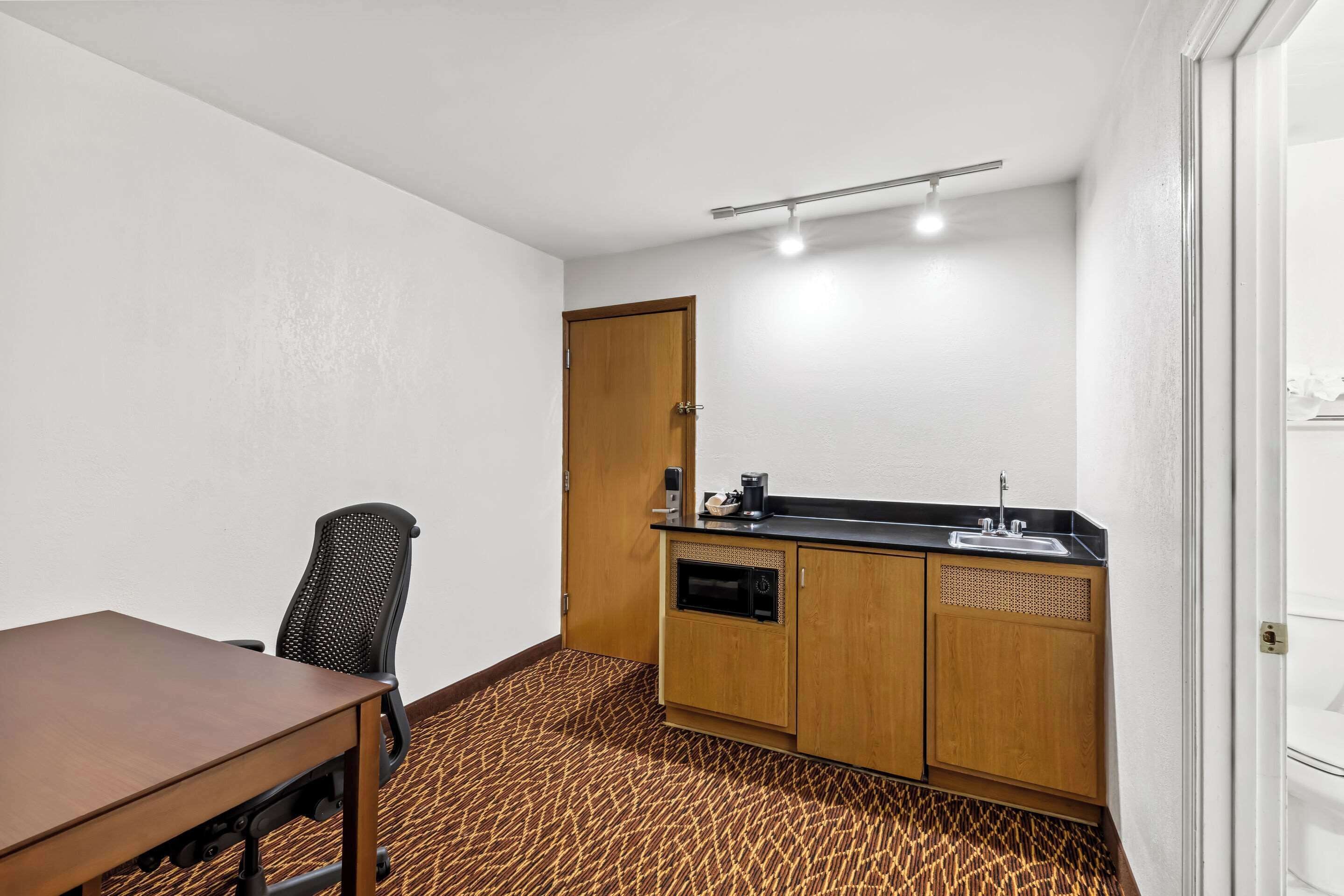 A small, compact room with a wooden table and a single office chair, a kitchenette with a sink and a microwave, and a patterned carpet.