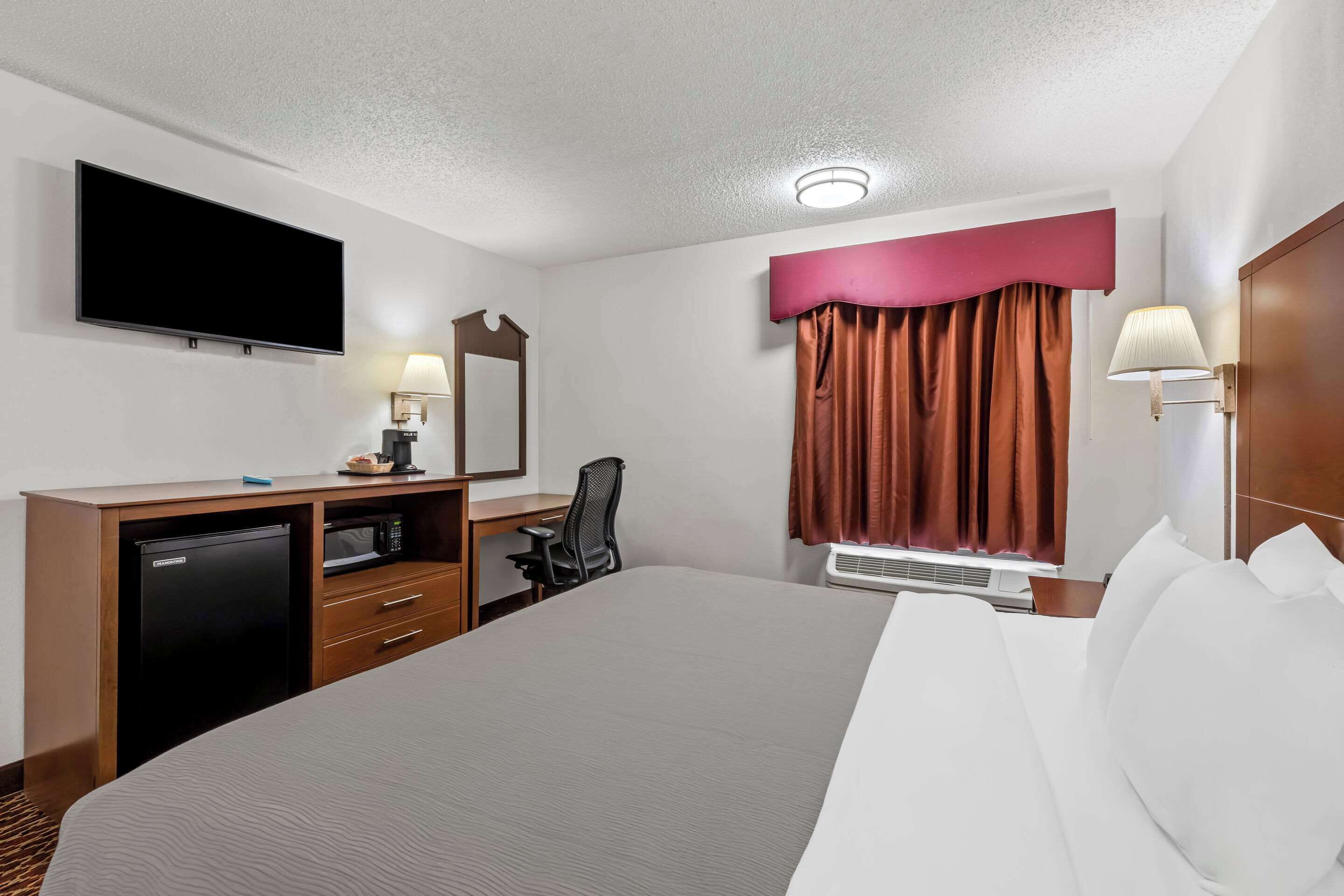 A hotel room with a large bed, a desk with a computer, a television, a microwave, and a window with curtains.