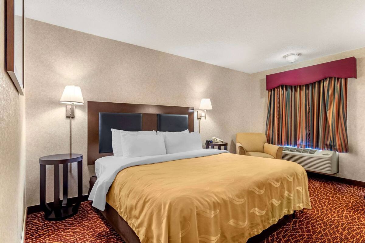 Une chambre d’hôtel comprenant un lit, deux lampes de chevet, une chaise, une fenêtre avec des rideaux et un appareil de climatisation.