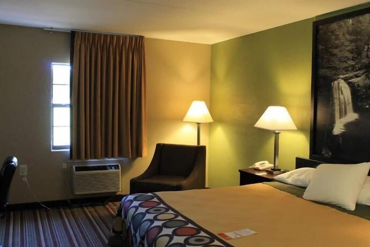 A hotel room with a bed, two bedside lamps, a chair, a desk with an air conditioner, and a large painting on the wall.