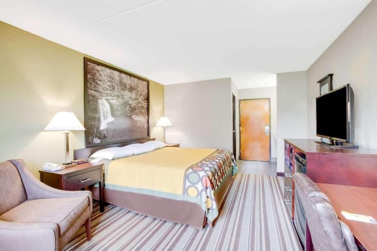 A hotel room with a bed, two bedside lamps, a TV, a sofa, a desk, and a painting on the wall.